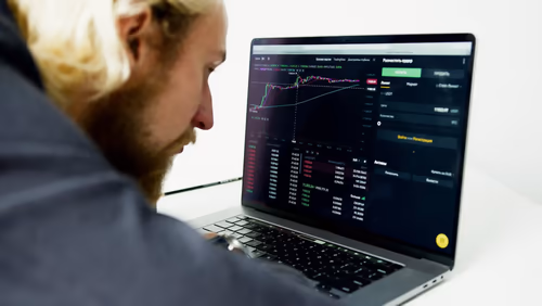 Over-the-shoulder view of a man at a laptop displaying a crypto trading dashboard